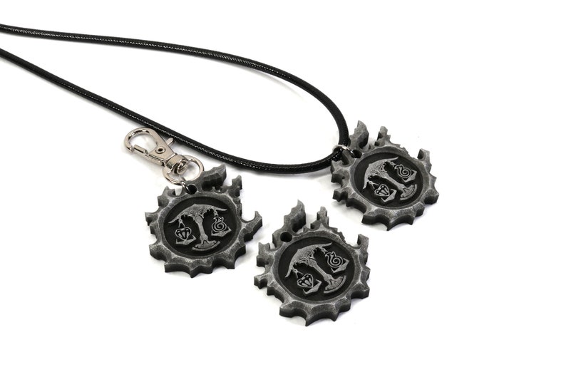 May include: A black leather necklace with a silver pendant featuring a stylized design. The pendant is a circular gear with a tree and scales in the center. The necklace comes with two additional pendants, one attached to a silver key chain.