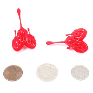 May include: Two red plastic heart-shaped objects with a dripping effect. The objects are on a white background. There are three coins below the objects.