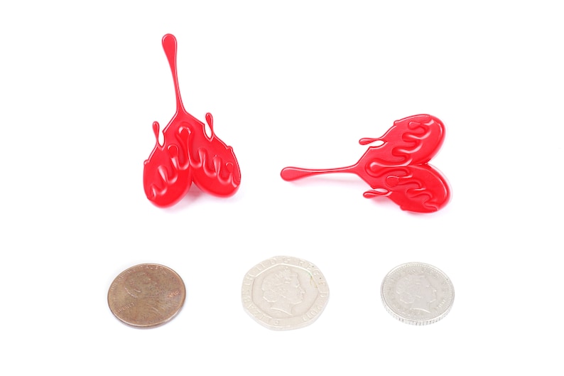 May include: Two red plastic heart-shaped objects with a dripping effect. The objects are on a white background. There are three coins below the objects.