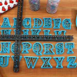 May include: A set of blue plastic alphabet cookie cutters on a wooden surface. The cutters are arranged in alphabetical order.