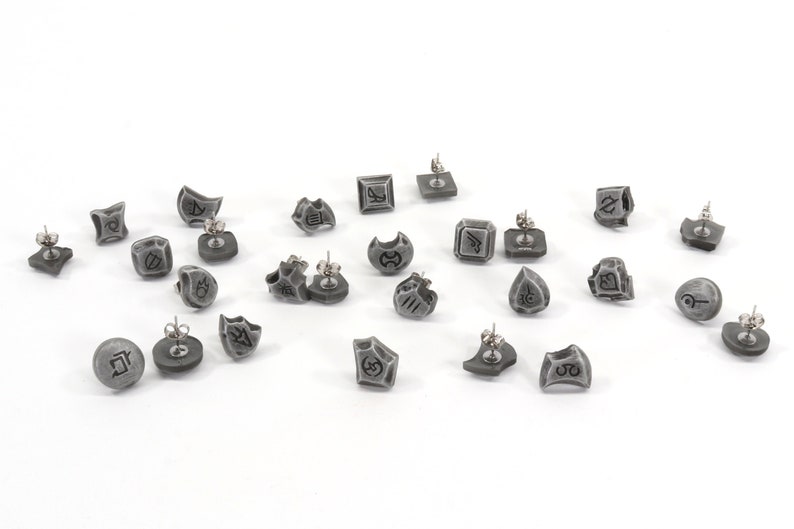 May include: A set of 18 gray earrings with different symbols and designs. The earrings are made of a metal alloy and have a silver finish.