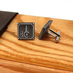 May include: A pair of silver cufflinks with a square design featuring a stylized sword and circle. The cufflinks are displayed on a wooden surface.
