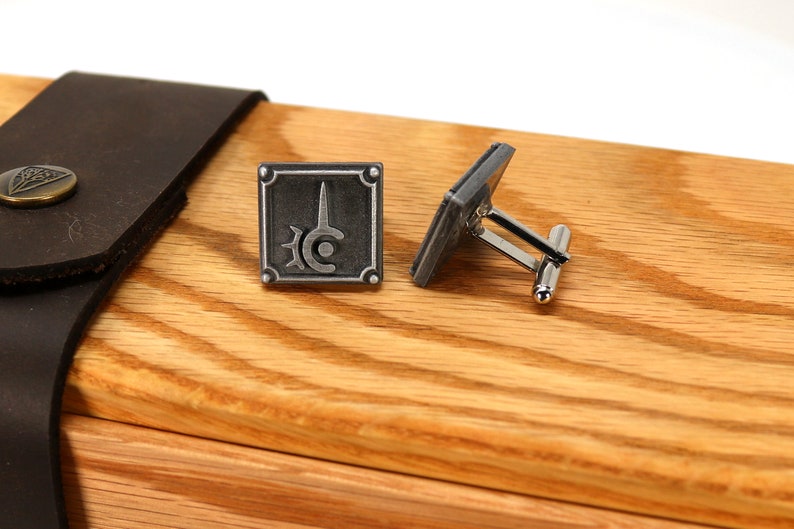 May include: A pair of silver cufflinks with a square design featuring a stylized sword and circle. The cufflinks are displayed on a wooden surface.