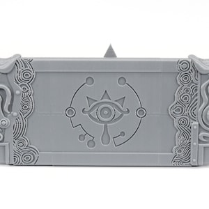 May include: Gray 3D printed Nintendo Switch case with a detailed design featuring the Triforce symbol and a stylized eye.
