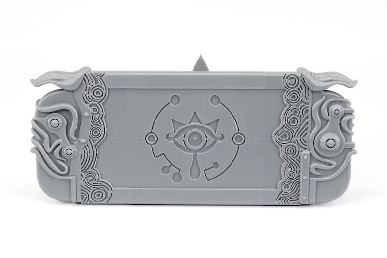 May include: Gray 3D printed Nintendo Switch case with a detailed design featuring the Triforce symbol and a stylized eye.