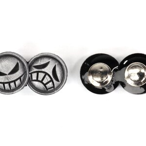 Op de afbeelding: Twee sets ronde zilveren en zwarte knoppen met een smiley-gezichtsontwerp. De zilveren knoppen hebben een fronsend gezicht met een opgeheven wenkbrauw en de zwarte knoppen hebben een glad oppervlak.