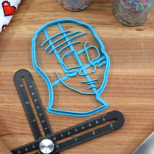 May include: A blue plastic cookie cutter shaped like a human head with lines across the face. The cutter is on a wooden surface.