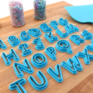 Op de afbeelding: Een set van 26 blauwe plastic alfabetkoekjesnijders op een houten oppervlak. De snijders zijn alfabetisch gerangschikt.