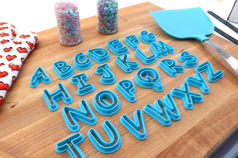 Op de afbeelding: Een set van 26 blauwe plastic alfabetkoekjesnijders op een houten oppervlak. De snijders zijn alfabetisch gerangschikt.