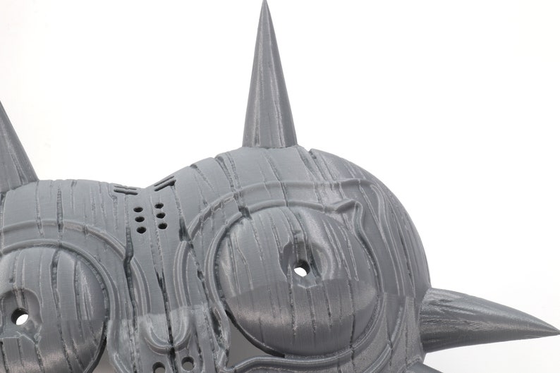 May include: A gray 3D printed mask with a sharp, pointed design. The mask has two large, round eyes and a series of small holes on the side.