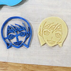 May include: A blue plastic cookie cutter and a yellow cookie in the shape of a person with short hair and a smiling face.