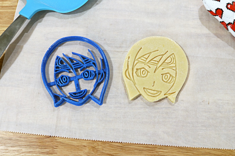May include: A blue plastic cookie cutter and a yellow cookie in the shape of a person with short hair and a smiling face.