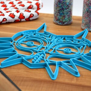 May include: A blue plastic cookie cutter shaped like the Majora's Mask from the Legend of Zelda video game series. The cutter is on a wooden surface.