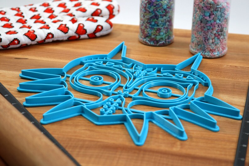 May include: A blue plastic cookie cutter shaped like the Majora's Mask from the Legend of Zelda video game series. The cutter is on a wooden surface.