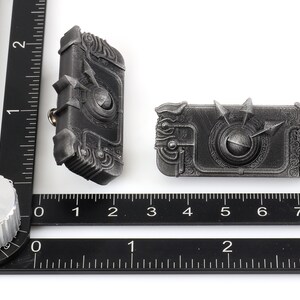 May include: Two silver-coloured metal pins with intricate designs. Each pin features a circular centre with a raised, textured surface and a surrounding border of detailed patterns. The pins are slightly different in size and shape.
