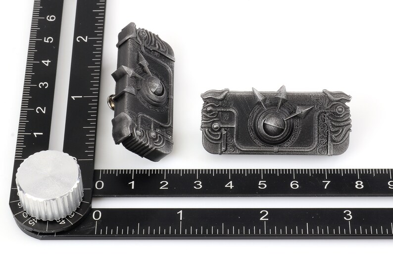 May include: Two silver-coloured metal pins with intricate designs. Each pin features a circular centre with a raised, textured surface and a surrounding border of detailed patterns. The pins are slightly different in size and shape.
