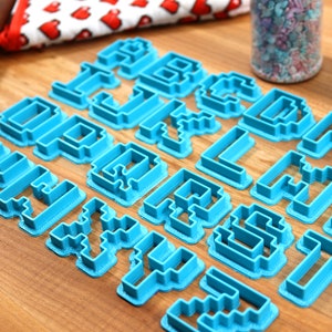 May include: A set of blue plastic cookie cutters shaped like letters of the alphabet in a pixelated style. The cutters are arranged on a wooden surface.