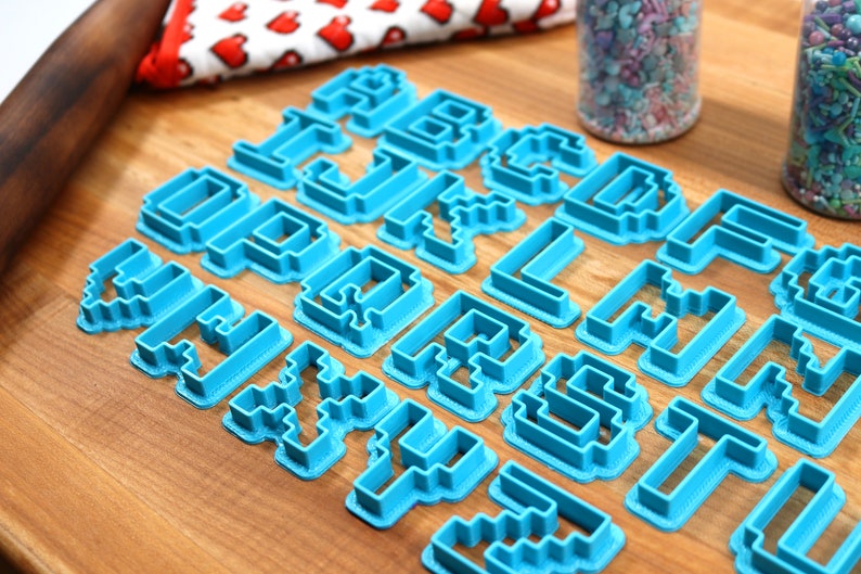 May include: A set of blue plastic cookie cutters shaped like letters of the alphabet in a pixelated style. The cutters are arranged on a wooden surface.