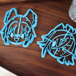 May include: Two blue plastic cookie cutters shaped like cartoon characters. The cutters are on a wooden surface.