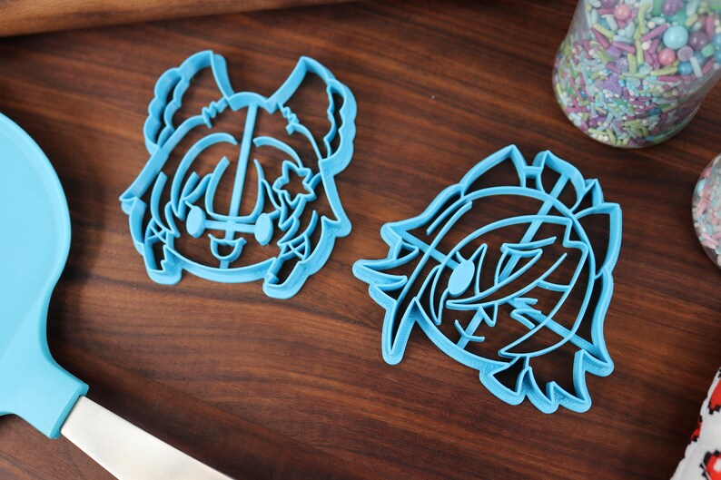 May include: Two blue plastic cookie cutters shaped like cartoon characters. The cutters are on a wooden surface.