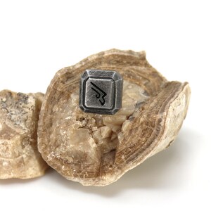 May include: A silver-colored metal charm with a stylized arrow design, resting on a piece of brown and white banded rock.