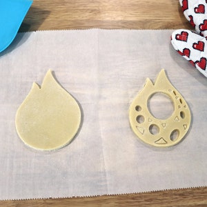 May include: Two unbaked cookie dough cutouts in the shape of a flame. One cutout is solid, while the other has a hole in the center and decorative cutouts around the edge.