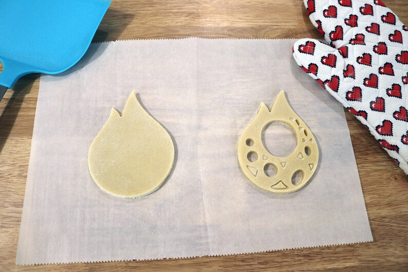 May include: Two unbaked cookie dough cutouts in the shape of a flame. One cutout is solid, while the other has a hole in the center and decorative cutouts around the edge.