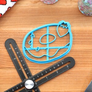 May include: A blue plastic donut cookie cutter with a strawberry on top. The cutter is on a wooden surface with a black ruler and a rolling pin.