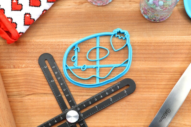 May include: A blue plastic donut cookie cutter with a strawberry on top. The cutter is on a wooden surface with a black ruler and a rolling pin.