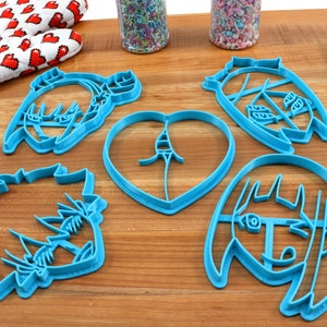 May include: Five blue plastic cookie cutters shaped like cartoon faces and a heart. The cutters are on a wooden surface.