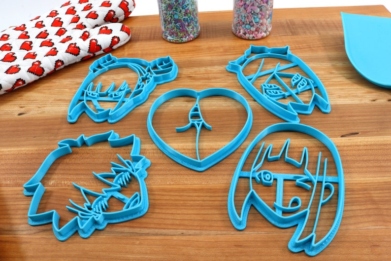 May include: Five blue plastic cookie cutters shaped like cartoon faces and a heart. The cutters are on a wooden surface.