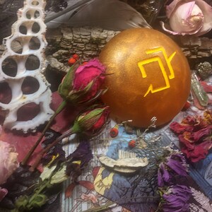 Puede incluir: Un huevo de color dorado con un s&iacute;mbolo amarillo en &eacute;l, rodeado de flores secas y otros elementos naturales.