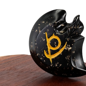 May include: Black obsidian crescent moon sculpture with a gold leaf finish and a yellow symbol on the surface. The sculpture is sitting on a wooden base.