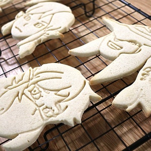 May include: Four plain sugar cookies decorated with icing designs of cartoon characters. The cookies are cooling on a black wire rack.