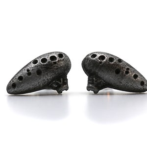 Pu&ograve; includere: Due piccoli ocarina grigio scuro, ciascuno con una serie di fori, poggiano su una superficie bianca. Gli ocarina hanno una forma a goccia e un aspetto strutturato, che suggerisce un materiale metallico o lapideo.