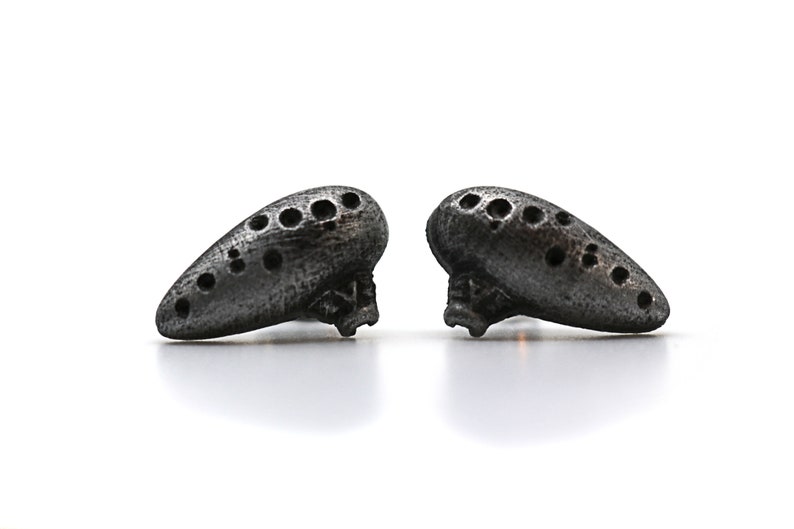 Pu&ograve; includere: Due piccoli ocarina grigio scuro, ciascuno con una serie di fori, poggiano su una superficie bianca. Gli ocarina hanno una forma a goccia e un aspetto strutturato, che suggerisce un materiale metallico o lapideo.