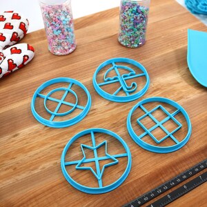May include: A collection of blue plastic cookie cutters in various shapes, including a star, umbrella, and geometric designs. The image also shows a heart-patterned oven mitt, sprinkles, and a blue spatula. A metal measuring cup and a ruler are also present.