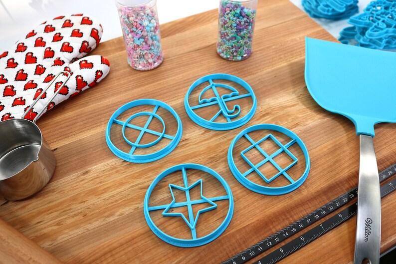 May include: A collection of blue plastic cookie cutters in various shapes, including a star, umbrella, and geometric designs. The image also shows a heart-patterned oven mitt, sprinkles, and a blue spatula. A metal measuring cup and a ruler are also present.