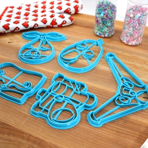 May include: A collection of blue plastic cookie cutters in various shapes, including a bow, an egg, and a bell, arranged on a wooden cutting board. A red and white oven mitt with heart patterns and two clear tubes filled with sprinkles are also visible.