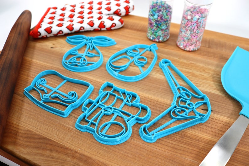 May include: A collection of blue plastic cookie cutters in various shapes, including a bow, an egg, and a bell, arranged on a wooden cutting board. A red and white oven mitt with heart patterns and two clear tubes filled with sprinkles are also visible.