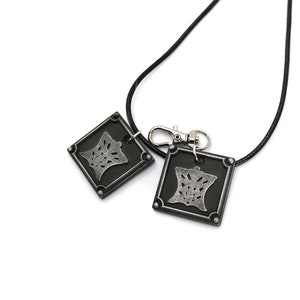 Puede incluir: Un collar negro con dos colgantes plateados. Cada colgante es un cuadrado con un dise&ntilde;o estilizado en su interior. El collar tiene un cierre plateado.
