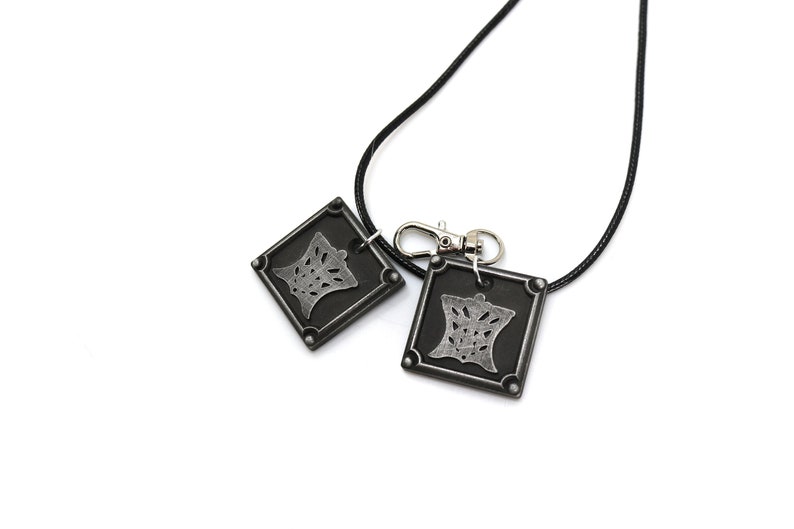 Puede incluir: Un collar negro con dos colgantes plateados. Cada colgante es un cuadrado con un dise&ntilde;o estilizado en su interior. El collar tiene un cierre plateado.