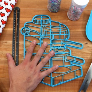 May include: Blue plastic cookie cutter shaped like an espresso machine. The cutter is large enough to fit a hand inside.