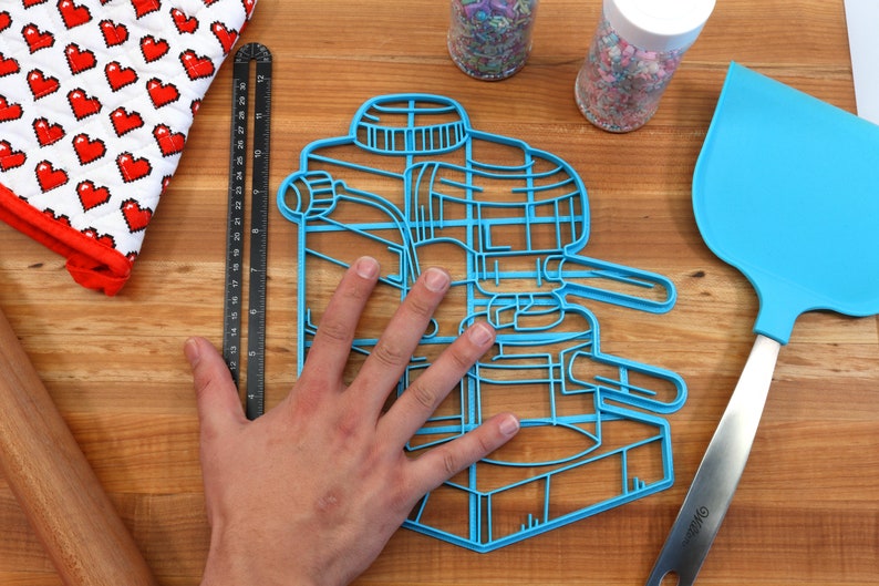 May include: Blue plastic cookie cutter shaped like an espresso machine. The cutter is large enough to fit a hand inside.