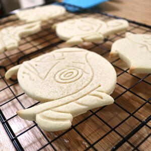 May include: Four plain sugar cookies, decorated with a variety of designs, are cooling on a black wire rack. The cookies are light brown in color and have a slightly rough texture.