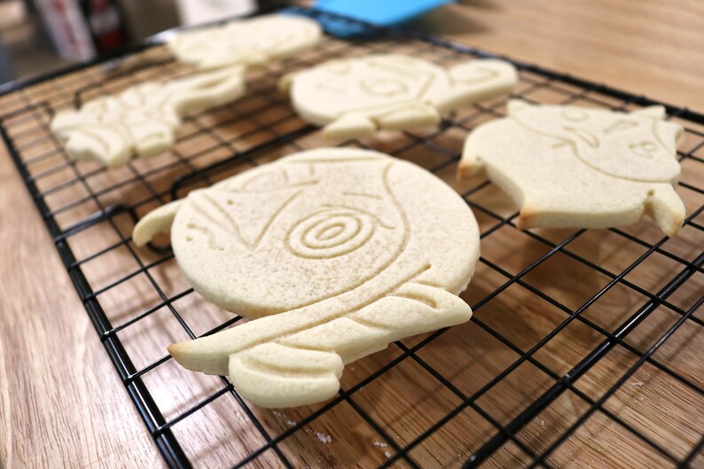 May include: Four plain sugar cookies, decorated with a variety of designs, are cooling on a black wire rack. The cookies are light brown in color and have a slightly rough texture.