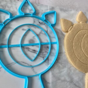 May include: A blue plastic cookie cutter shaped like a sun with a large circle in the center and a smaller circle inside the larger circle. The cookie cutter is next to a cookie that has been cut out using the cookie cutter.