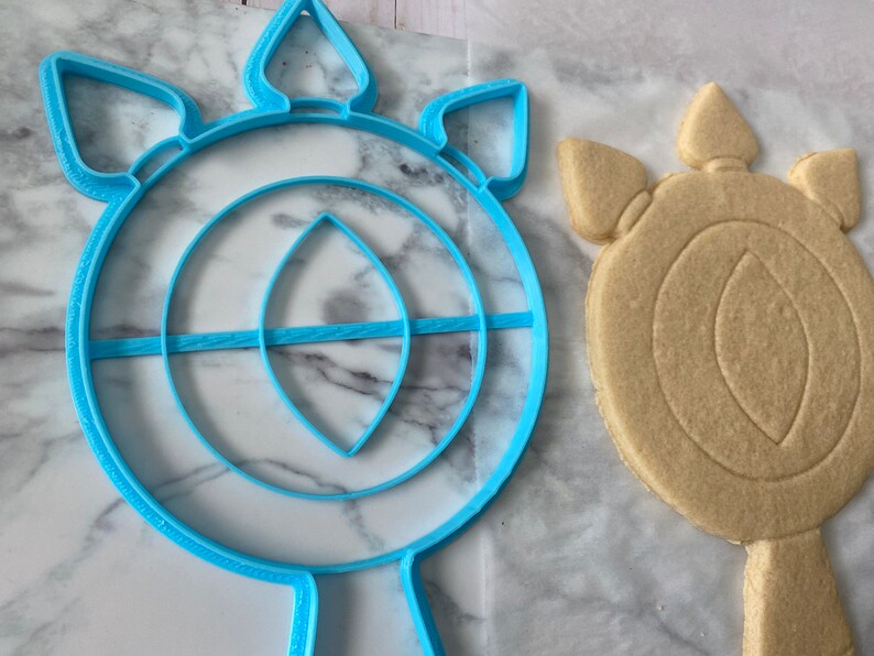 May include: A blue plastic cookie cutter shaped like a sun with a large circle in the center and a smaller circle inside the larger circle. The cookie cutter is next to a cookie that has been cut out using the cookie cutter.