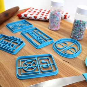 May include: A set of five blue plastic cookie cutters shaped like a cassette tape, a record player, and a music note. The cutters are on a wooden surface with sprinkles in jars and a rolling pin.
