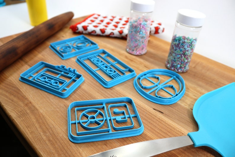 May include: A set of five blue plastic cookie cutters shaped like a cassette tape, a record player, and a music note. The cutters are on a wooden surface with sprinkles in jars and a rolling pin.